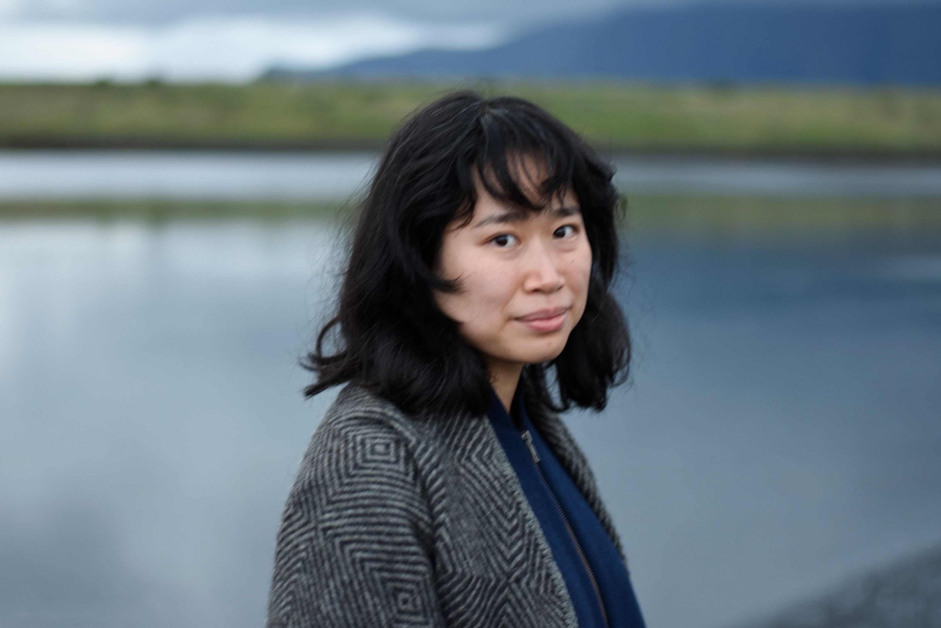 Image of Studio Director Luisa Ji standing by the water wearing a gray jacket and maybe, sort-of smiling.