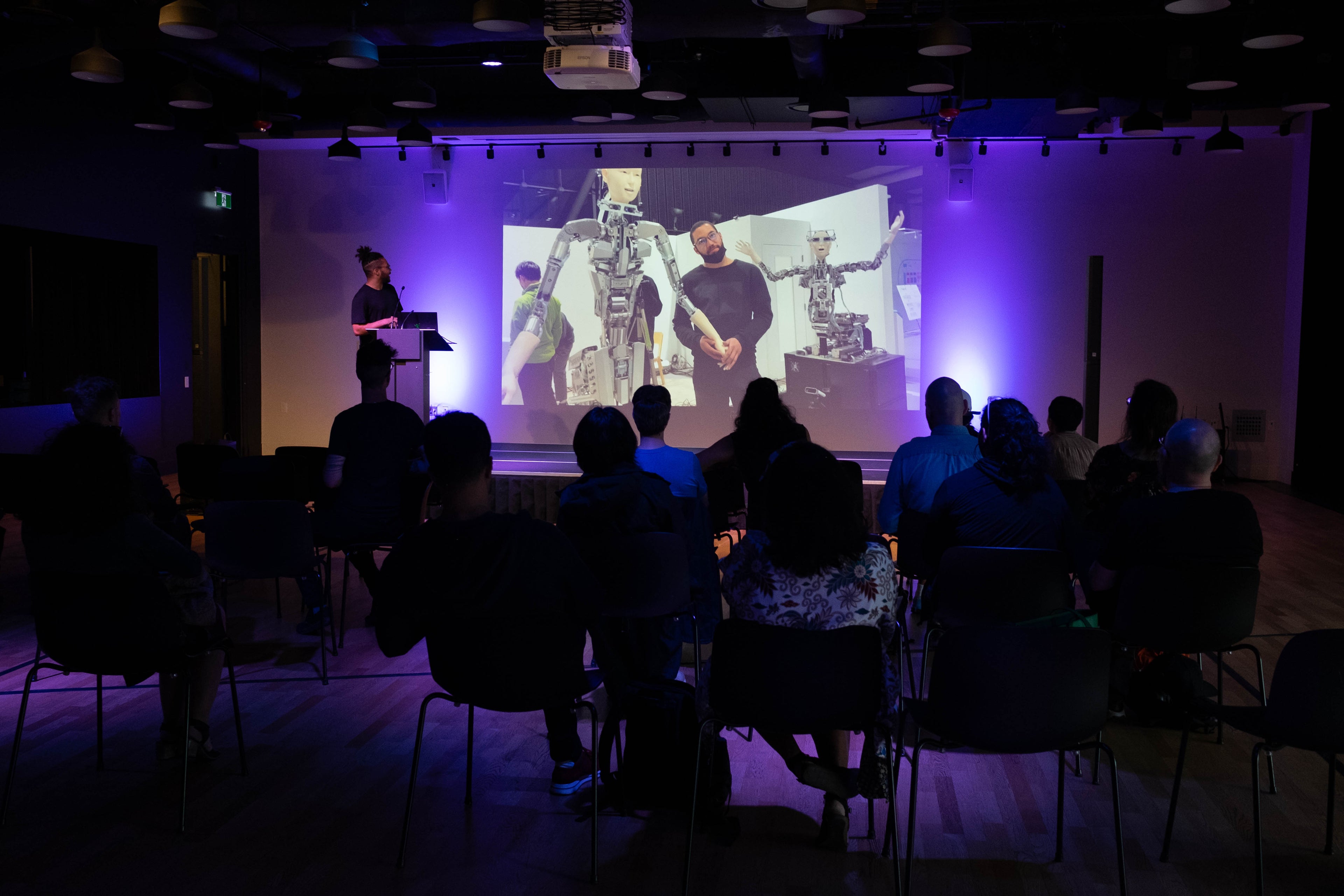 Photograph of artist Maurice Jones standing in front of a slide where is holding a robot's hand. The picture is from Artscape Launchpad from the Carnival of Algorithmic Culture in 2023.