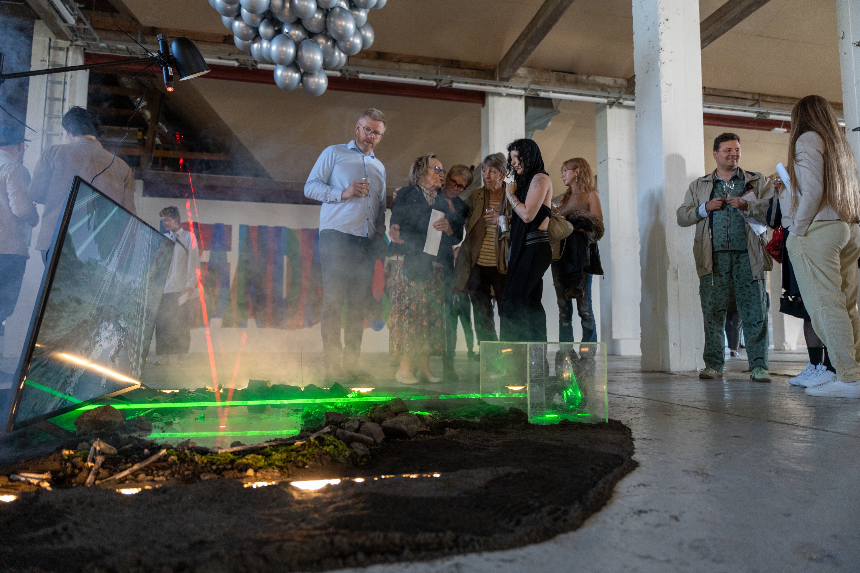 Photograph taken from ground level of a crowd of various generations gathered around an art exhibit at SIM in Iceland. The art has a screen with a digital representation of a river with multipole green and red lasers simulating nuclear strikes.