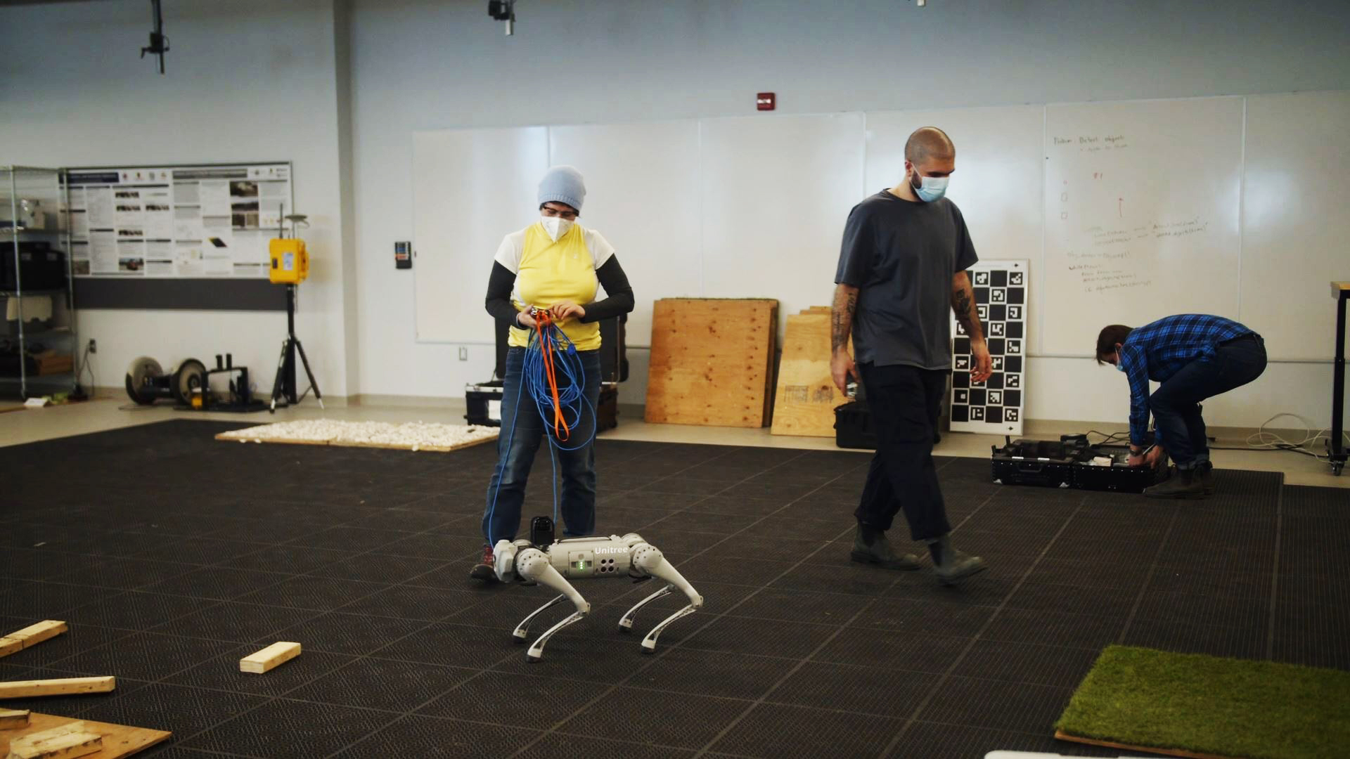 Two Ferment AI resident artists standing on a rubber mat floor. One, female, is holding a remote and controlling a small robotic dog.